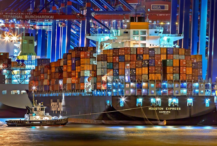 Es ist ein riesiges, voll beleuchtest Containerschiff zu sehen im Hafen angelegt. Auch ein Schlepper ist zu sehen.