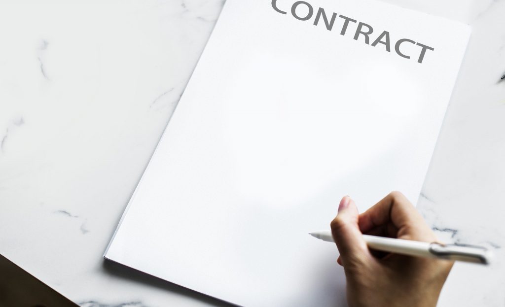 Es liegt ein Stapel Papiere auf einem weißen Tisch, auf dem obersten Papier steht "Contract" außerdem ist eine Hand mit einem Stift in der Hand zu sehen